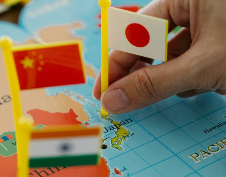 A hand holding the Japanese flag pinpoints Japan on a world map alongside China and India flags.